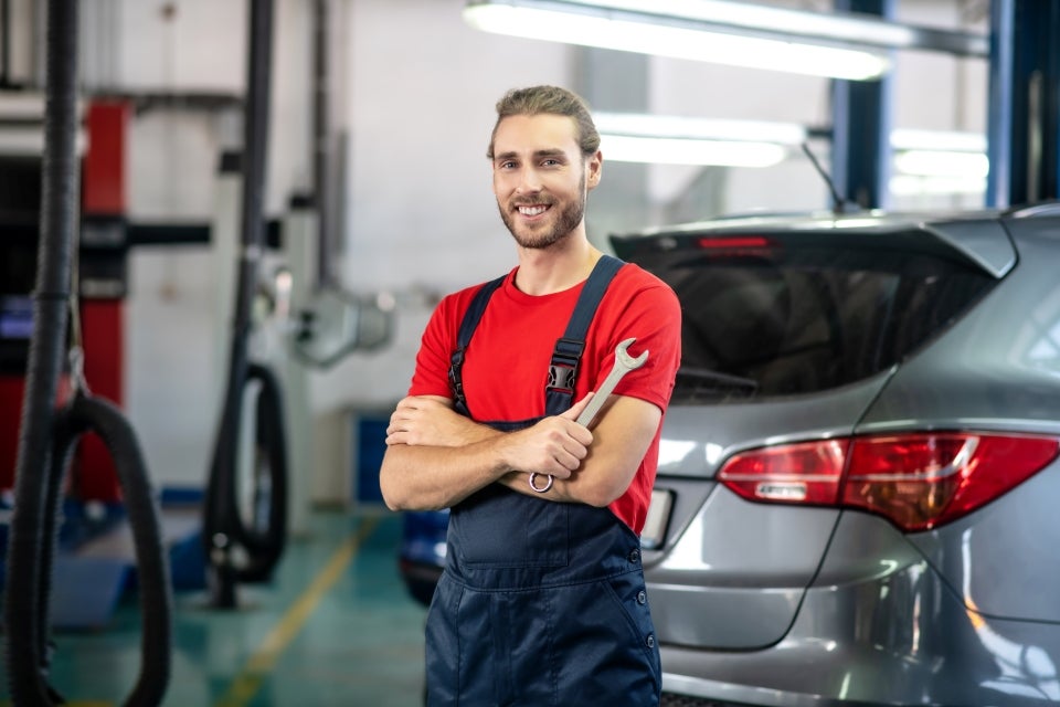 A GMC Service Center certified professional mechanic is wearing a red t-shirt with dark blue overalls is holding a wrench with folded arms and is smiling at us in front of a grey SUV in the shop.